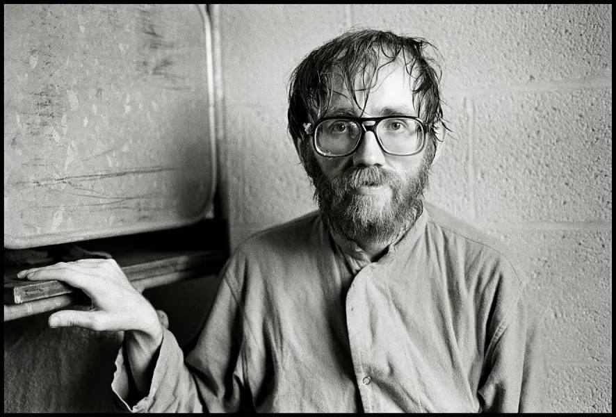 Schwarzweißfoto eines bärtigen Mannes mit Brille in einem Hemd, der vor einer Wand mit Papieren oder Dokumenten sitzt und in die Kamera blickt.