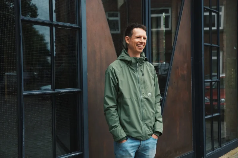 Andreas in einer grünen Jacke und Jeans steht lächelnd vor einer braunen Tür mit schwarzen Fenstern. Er hat die Hände in den Taschen und schaut nach links. Der Hintergrund zeigt eine moderne Fassade mit dunklen Fensterrahmen.