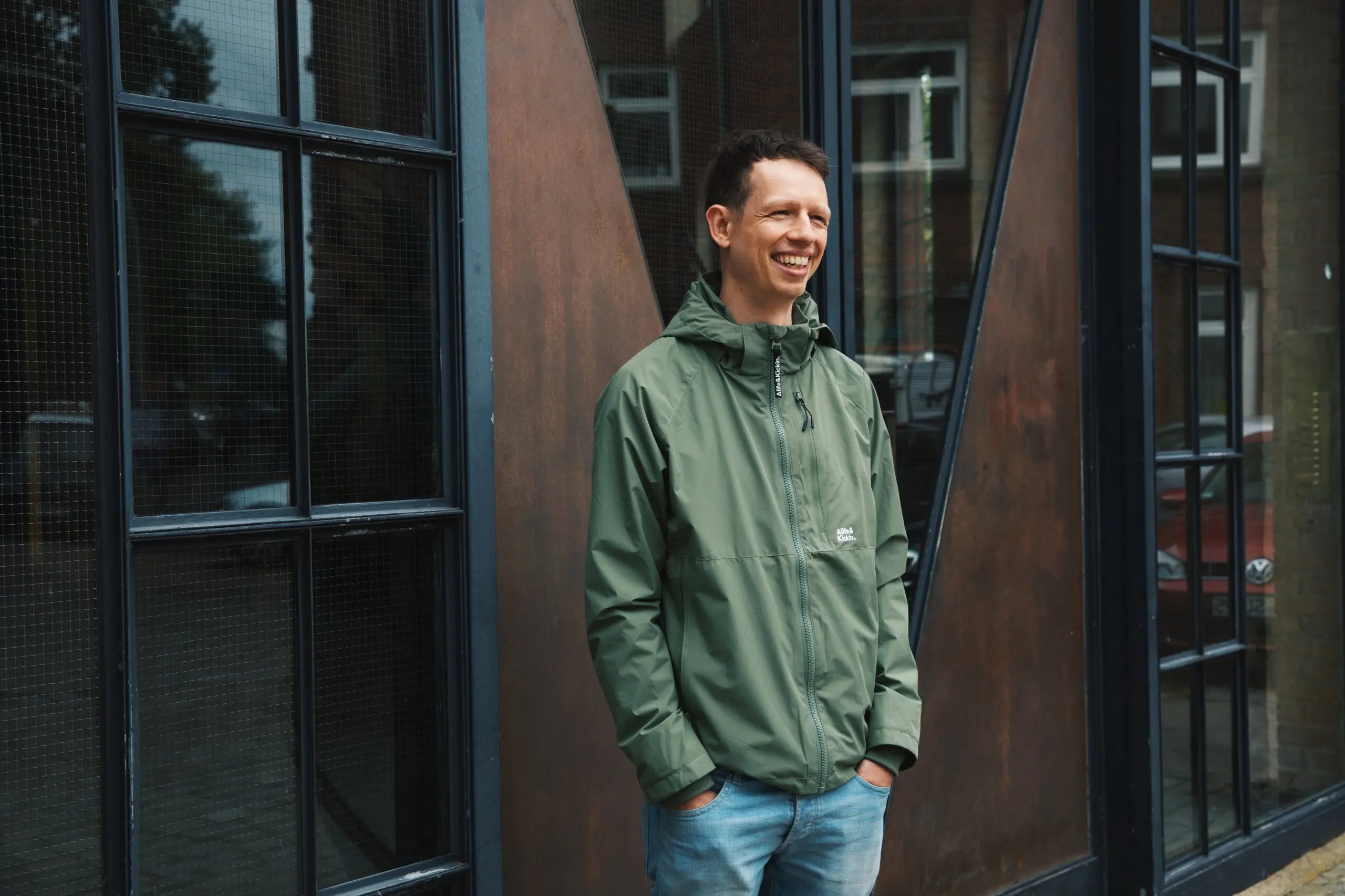 Andreas in einer grünen Jacke und Jeans steht lächelnd vor einer braunen Tür mit schwarzen Fenstern. Er hat die Hände in den Taschen und schaut nach links. Der Hintergrund zeigt eine moderne Fassade mit dunklen Fensterrahmen.